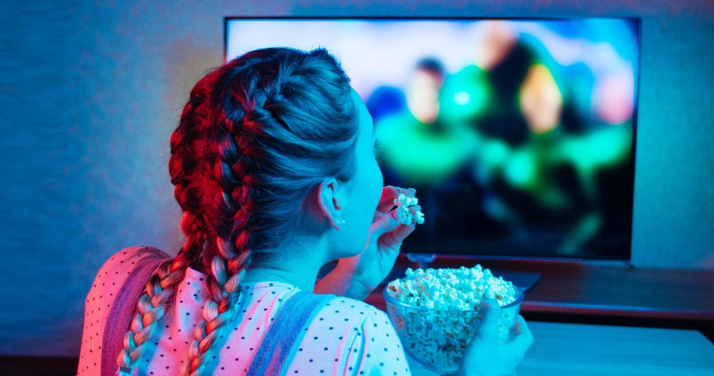 دختری با موهای بافته رو به تلویزیون نشسته و فیلم نگاه می‌کند و پاپ‌کُرن می‌خوردد