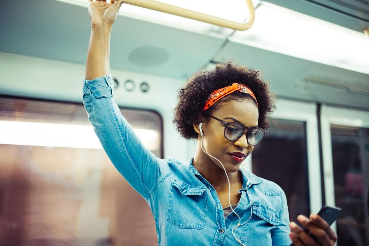 دختری رنگین پوست در اتوبوس ایستاده با موهای فر و عینک که درحال گوش دادن به موسیقی با هندزفری است